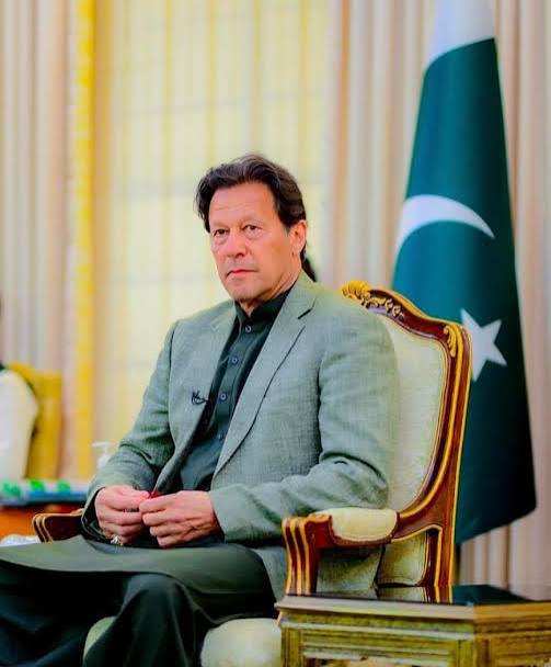 Imran Khan seated in an armchair in a grey blazer, with the Pakistan flag behind him