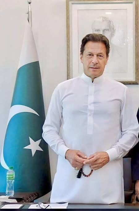 Imran Khan in a white shalwar kameez standing beside the Pakistan flag